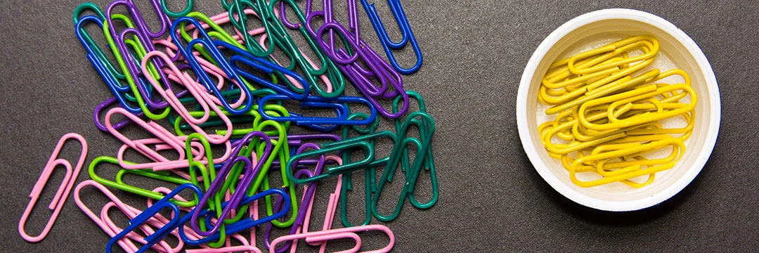 Yellow paper clips in a white bowl and many colorful paper clips lying jumbled on the table