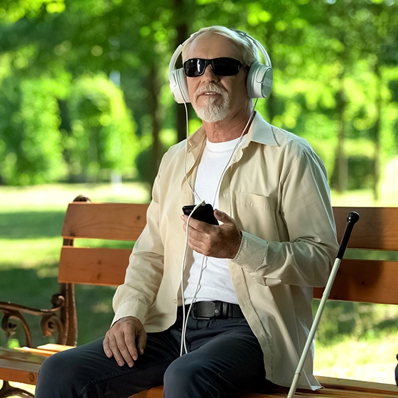 Ein älterer blinder Mann sitzt auf der Parkbank hat sein Handy in der Hand und trägt Kopfhörer 