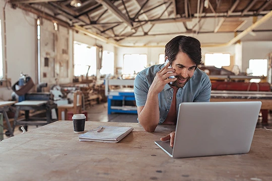 Ein Mann in seiner Werkstatt vor dem Laptop mit dem Handy am Ohr