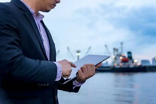 Ein Mann steht im Anzug im Hafen und schaut auf seinem Tablet etwas nach