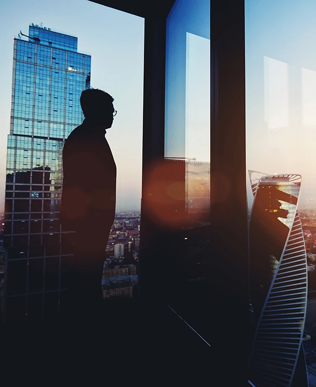 Geschäftsmann steht in seinem Büro mit Glasfront und schaut sich den Sonnenuntergang an