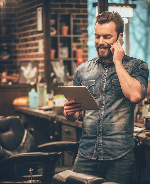 Cheerful young bearded man talking on mobile phone and looking at digital tablet while standing at barbershop