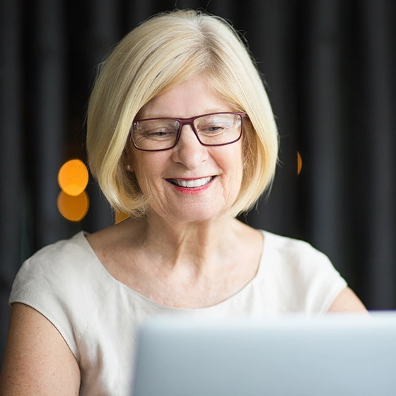 Ältere Frau mit blonden Haaren und Brille sitzt vor dem Laptop