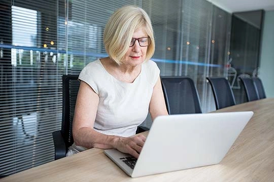 ältere Dame sitzt in einem Meetingraum und arbeitet auf einem Laptop