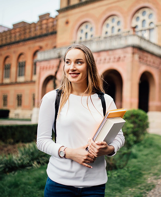 Eine junge Frau mit langen Haaren steht mit Büchern in der Hand vor einem großen Backsteingebäude