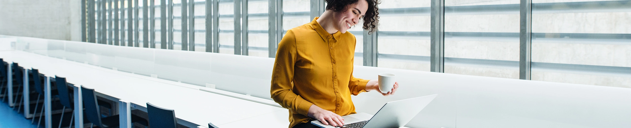 Eine Frau mit gelber Bluse sitzt auf einem Tisch und schaut auf ihren Laptop