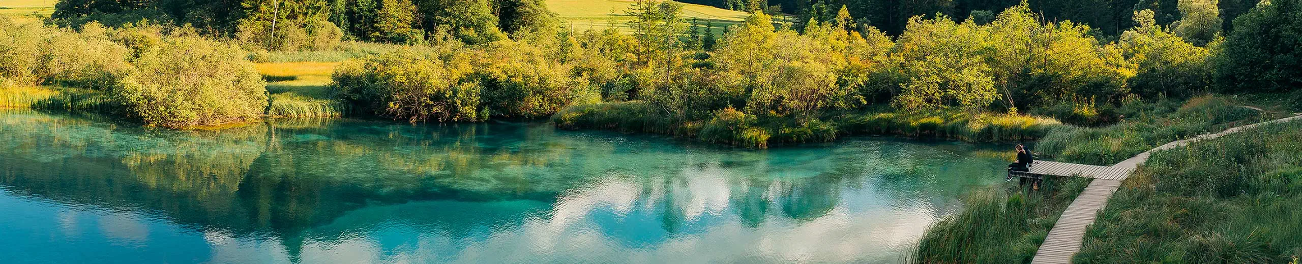 Ein See, mit Büschen und einem Steg, auf dem 2 Personen sitzen