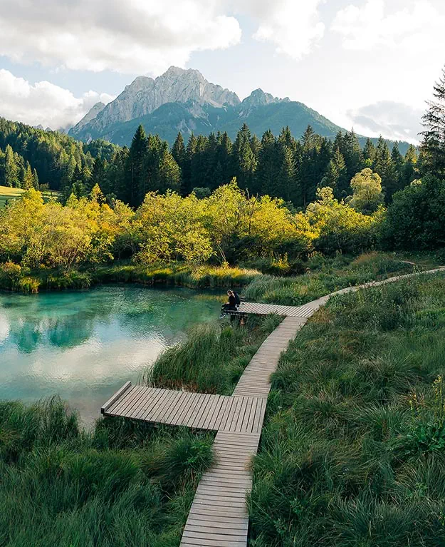 Ein See, mit Büschen und einem Steg, auf dem 2 Personen sitzen