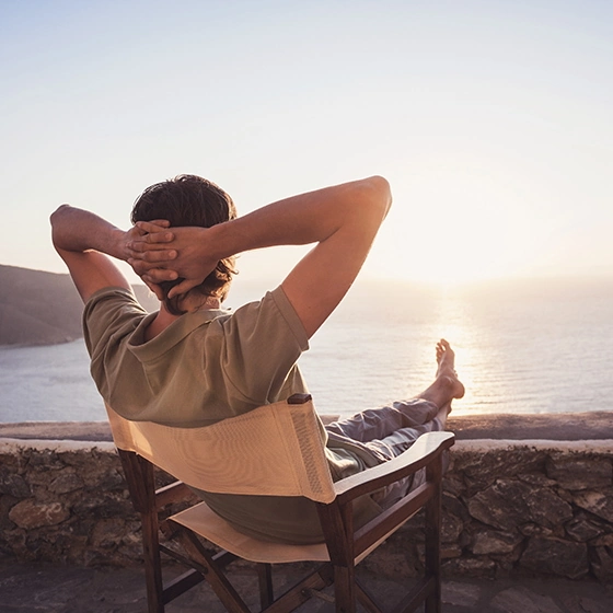 Ein Mann sitzt auf einer Terasse über dem Meer und schaut in die untergehende Sonne