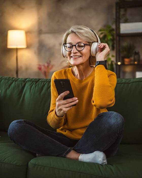 Eine Frau sitzt mit Kopfhörern auf der Couch und schaut aufs Handy