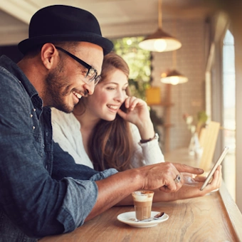 Ein junger Mann und eine junge Frau sitzen in einem Kaffeehaus und schauen sich etwas auf einem Smartphone an.