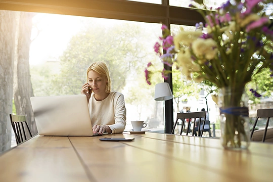 Eine Frau sitzt mit ihrem Handy am Ohr vor dem Laptop an einem Holz Esstisch