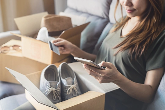 Eine Frau mit einem geöffneten Paket, in dem Schuhe sind, auf dem Schoß und der Kredikarte in der Hand