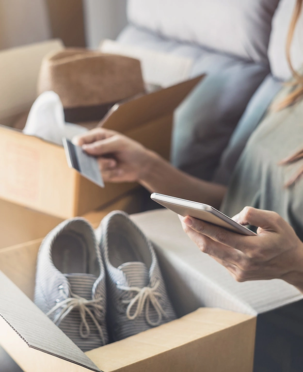 Eine junge Frau sitzt auf einem Sofa mit einem Schuhkarton auf dem Schoß, einer Kreditkarte in der linken und einem Smartphone in der rechten Hand