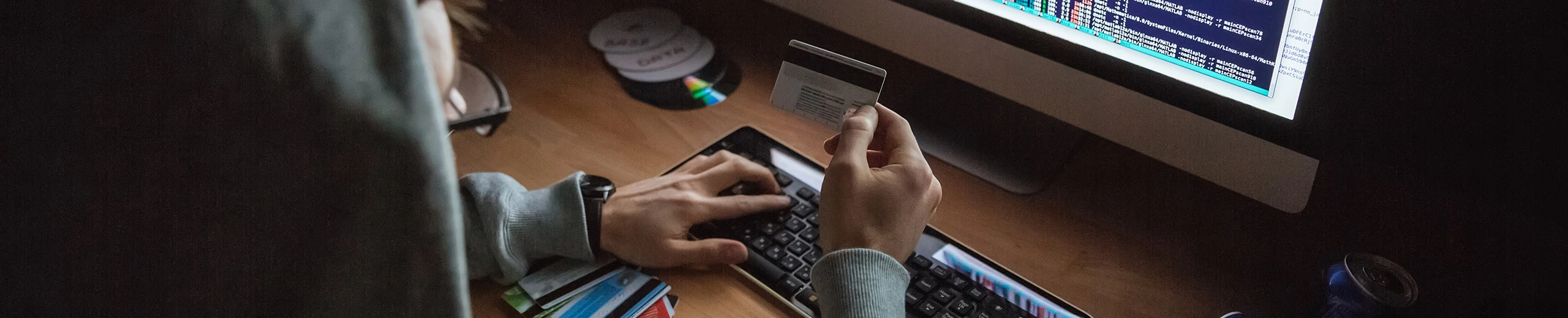 Ein Mann sitzt vor einem Computer tippt mit einer Hand auf die Tastatur und mit der anderen Hand hält er eine Kreditkarte