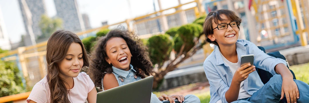 3 Kinder sitzen mit Laptop und Handy in der Wiese mitten in der Stadt