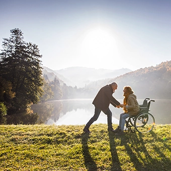 Ein Paar vor einem See in der Sonne, die Frau sitzt im Rollstuhl, der Mann steht ihr gegenüber